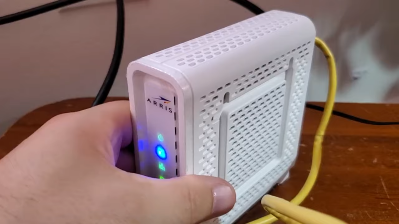 The Image Shows a Person Holding an Arris Modem with Several Indicator Lights On, Connected via A Yellow Ethernet Cable